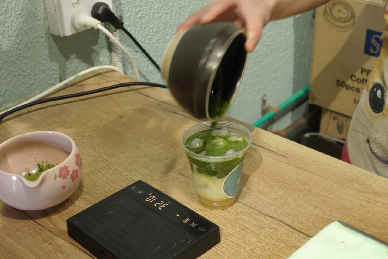 A person's hand pours green powder into a plastic cup to make a matcha drink.