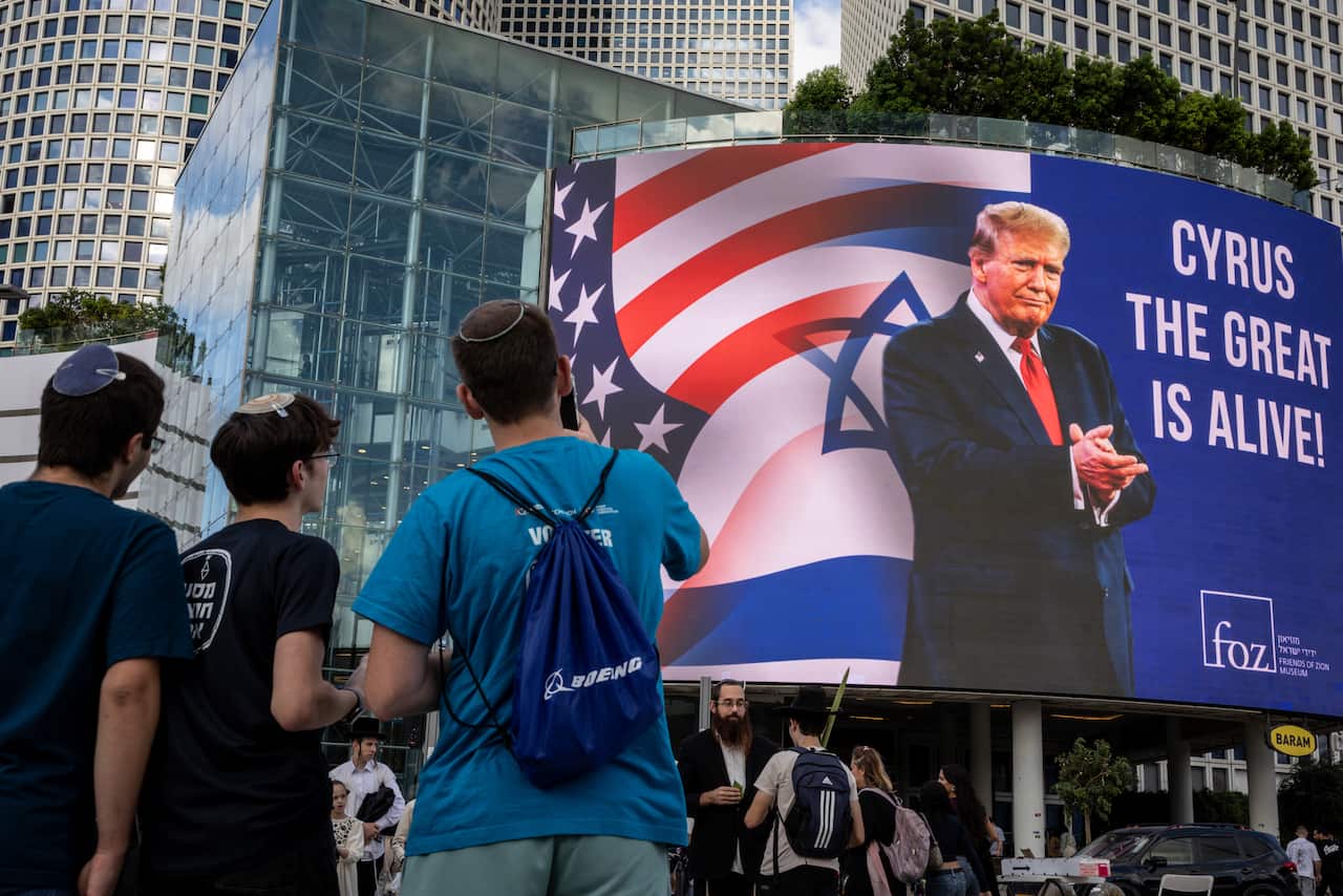 A group of three young men wearing kippahs look at a large billboard showing a picture of Donald Trump and the words "Cyrus the Great is alive!"