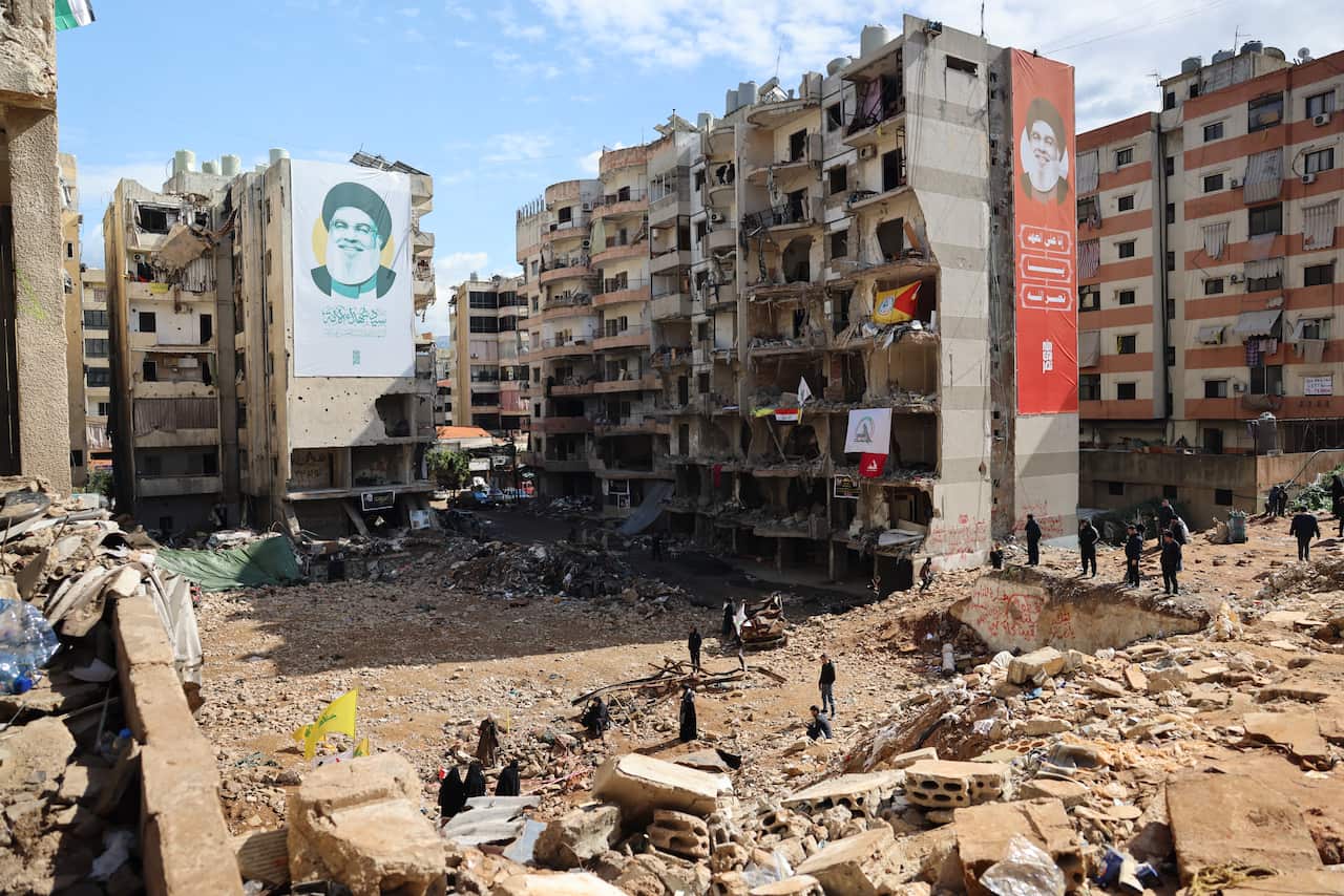 A residential area in ruins after a bombing, with damaged multi-storey buildings and a few people standing on the rubble. Two posters of a grey-bearded older man smiling hang on the sides of the damaged buildings