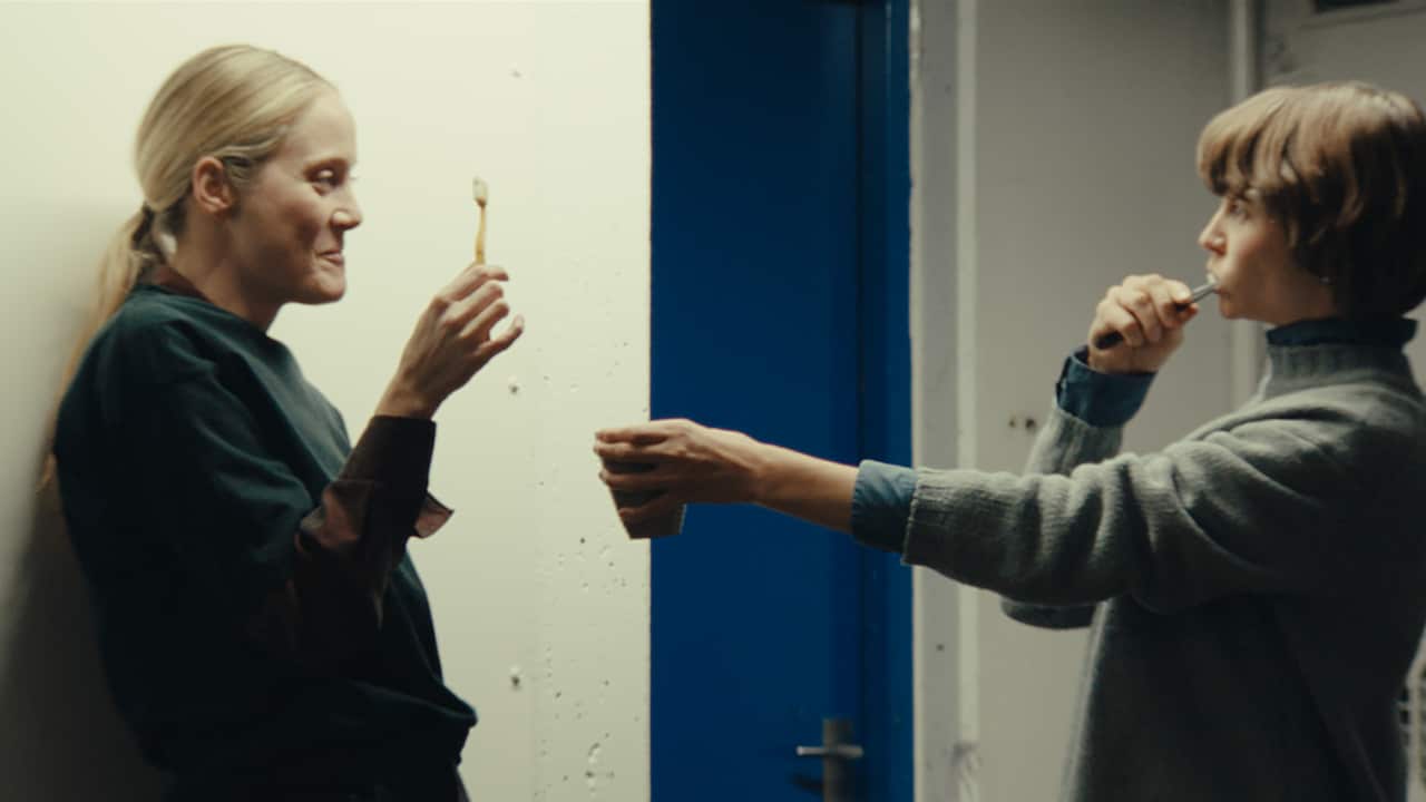 Two women stand in front of a white wall and a bright blue door. They are laughing as they both clean their teeth. 