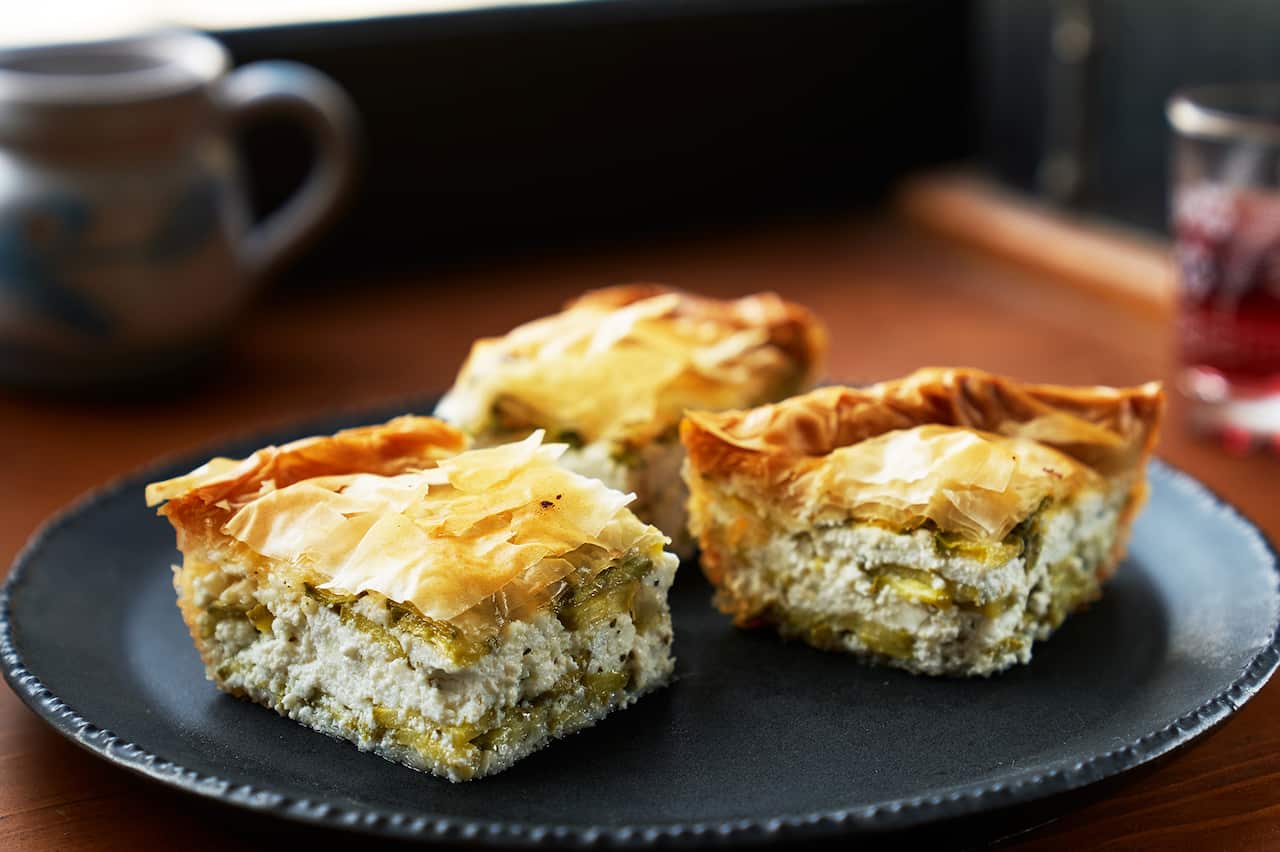 Three pieces of pie, with a cream filling studded with green and flaky golden filo pastry o top, sit on a dark round plate on a table. A mug and glass can be see, out of focus, in the background. 