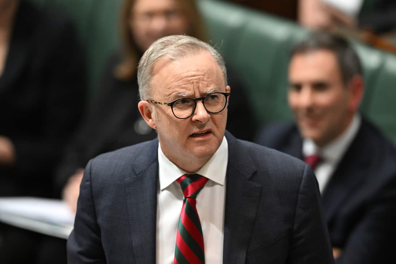 A man in a dark suit, white collared shirt, and diagonally striped red and green tie looks forward with a serious expression on his face.