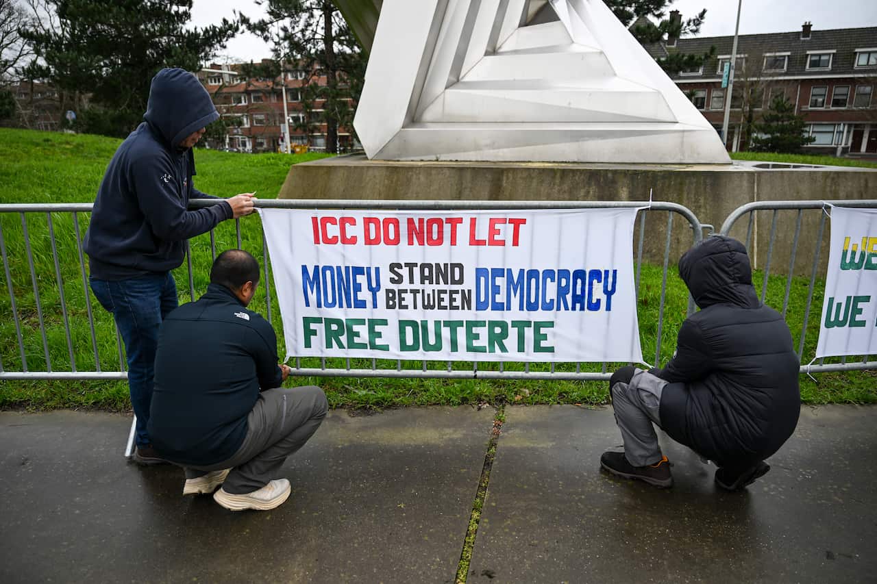 A group of people in black jackets attaches a protest sign to a temporary metal fence. The fence surrounds a triangular statue. The text of the poster reads: ICC DO NOT LET MONEY STAND BETWEN DEMOCRACY, FREE DUTERTE.