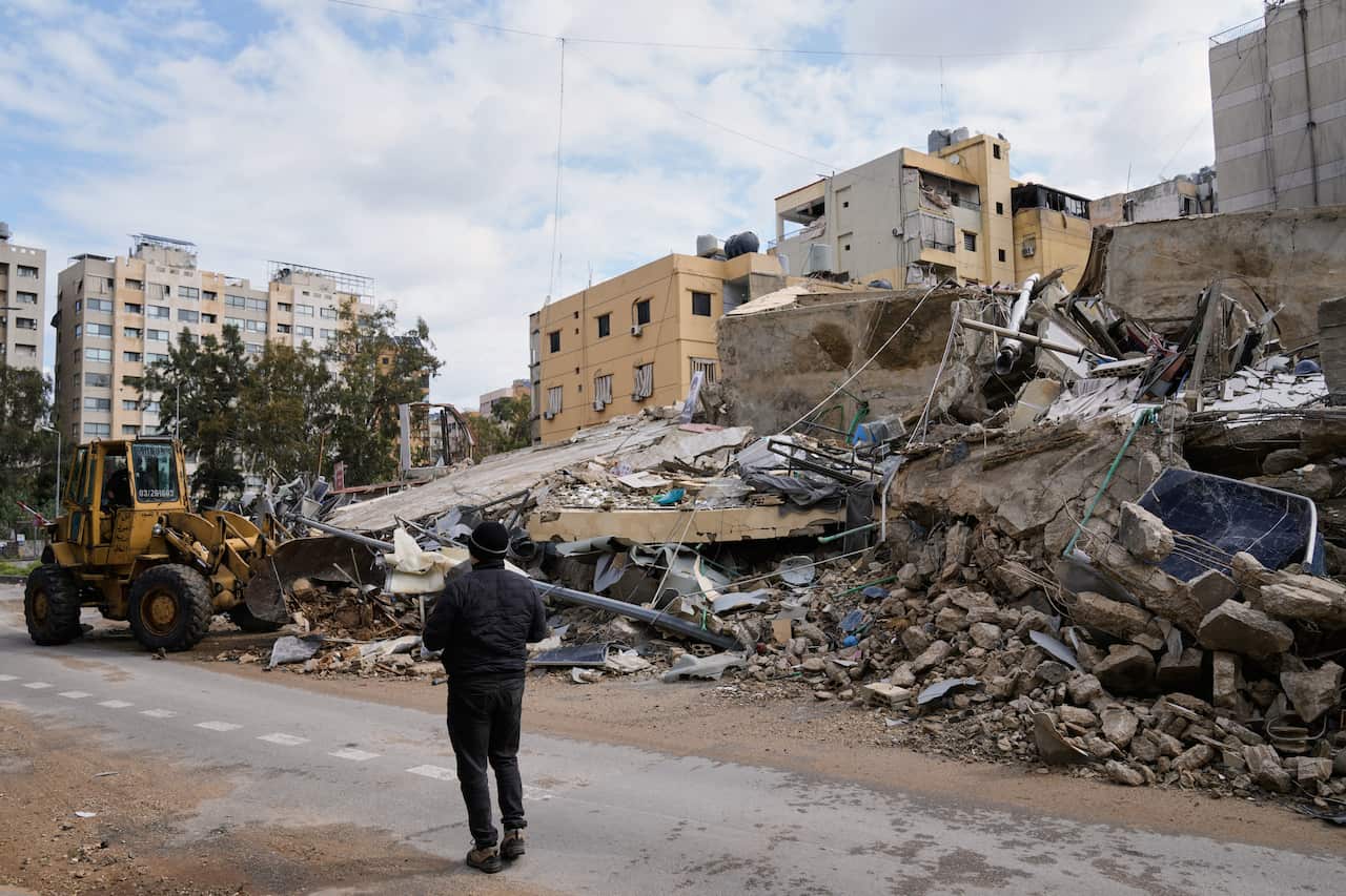 A person standing on the road in front of the rubble of a heavily damaged building.