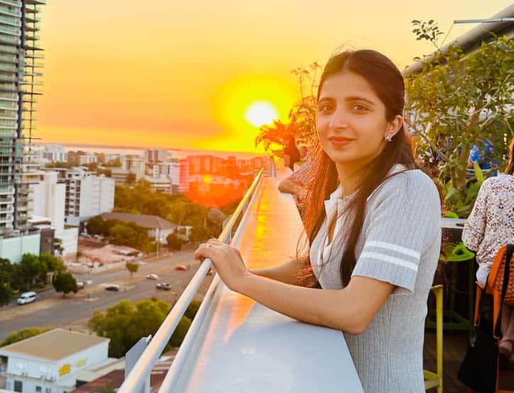 A woman standing on a balcony with her arms on a barrier. The sunset can be seen behind her.