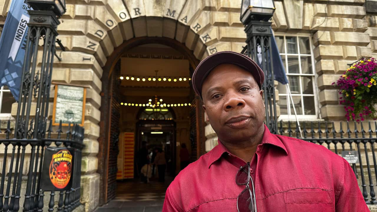 A Black man wearing a red shirt and a flat cap looks into the camera, posing in front of a building entrance with 'Indoor market' written above it