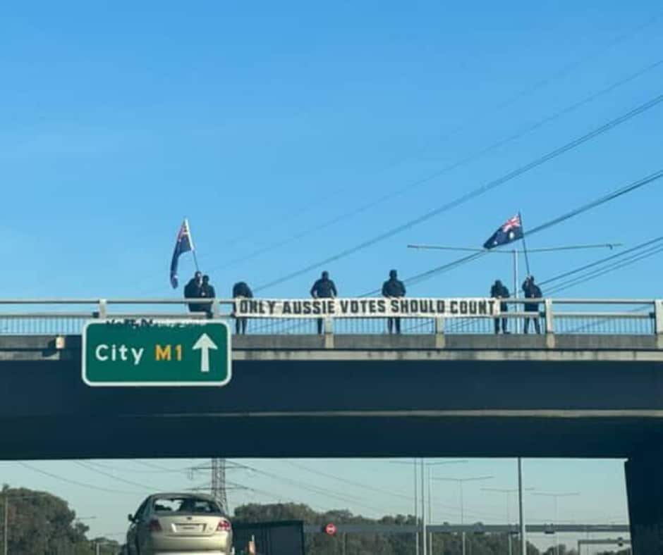 A group dressed in black, holding Australian flags and a "ONLY AUSSIE VOTES SHOULD COUNT" banner on a bridge