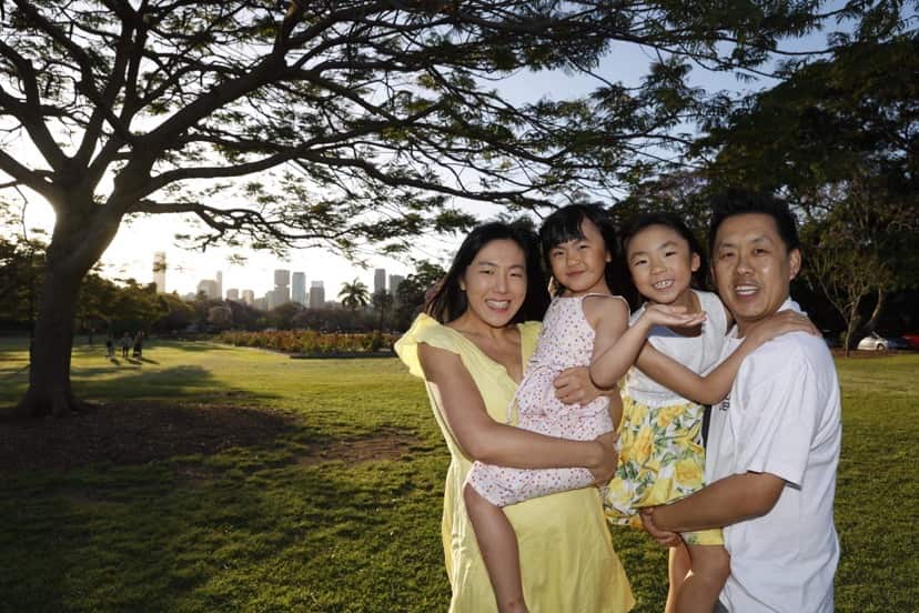 A mum, dad and two young girls smile and pose in a park with the cityscape in the background.