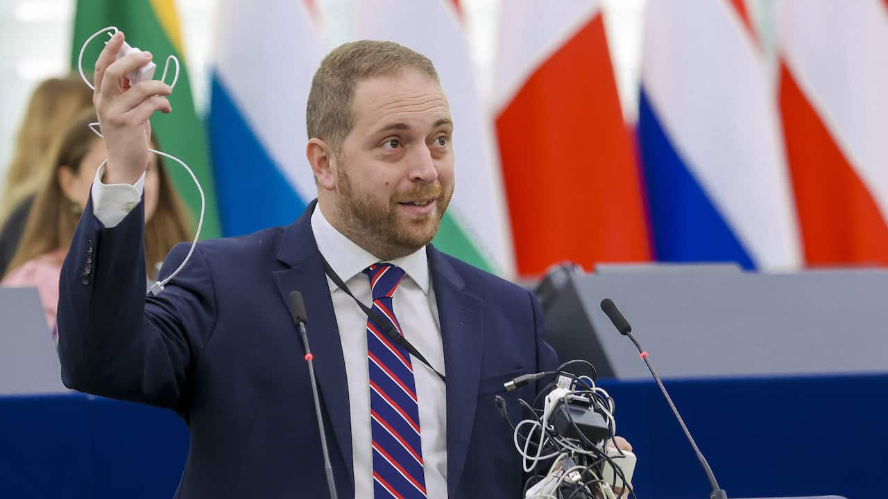 Alex Agius Saliba holds up a handful of chargers and cords while speaking into a microphone.