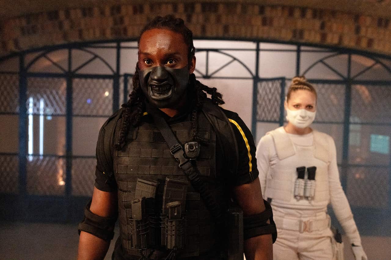 A man in dark combat gear and a black half mask stands in a building. A woman in white protective hear and a white mask stands to one side behind him.