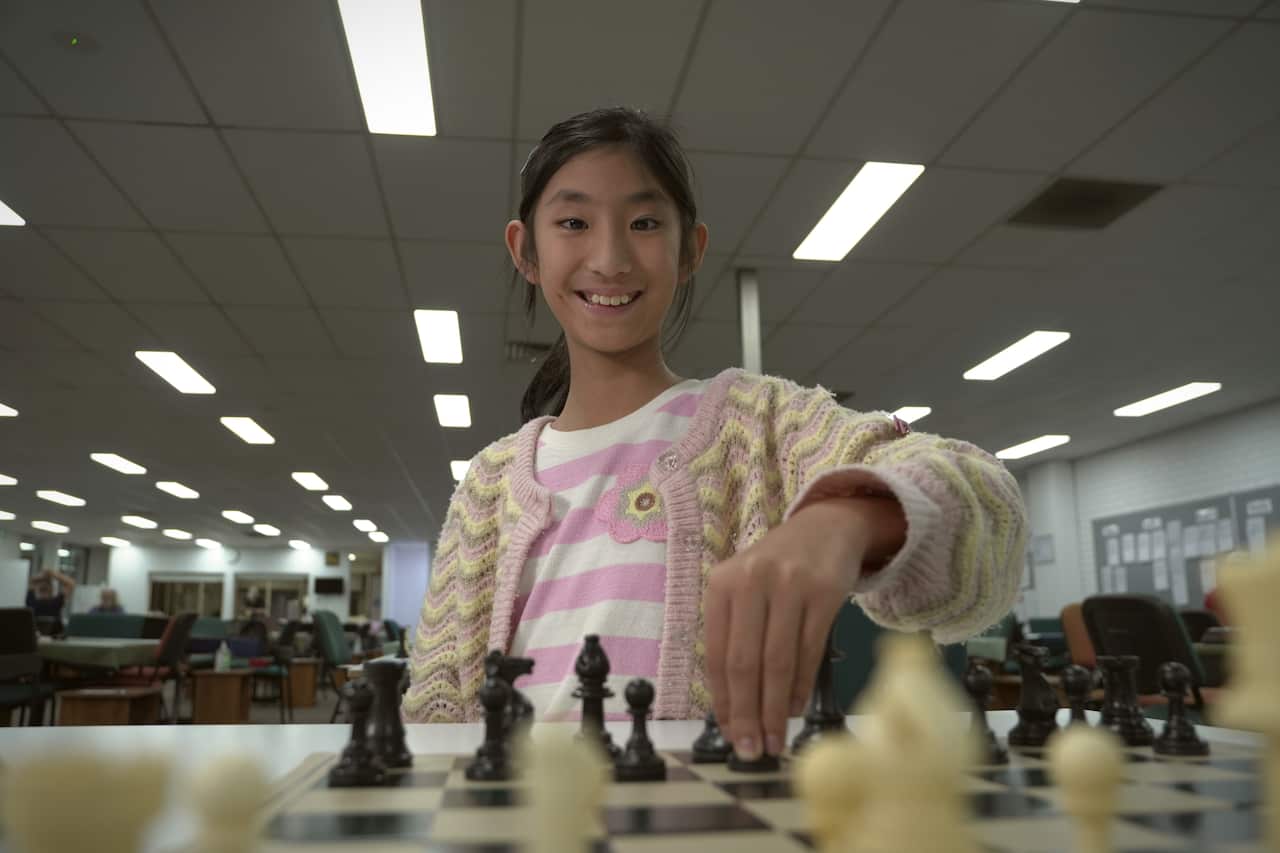 A young girl wearing a cardigan over a striped top is playing chess. She is smiling and looking at the chessboard as she moves a piece around.