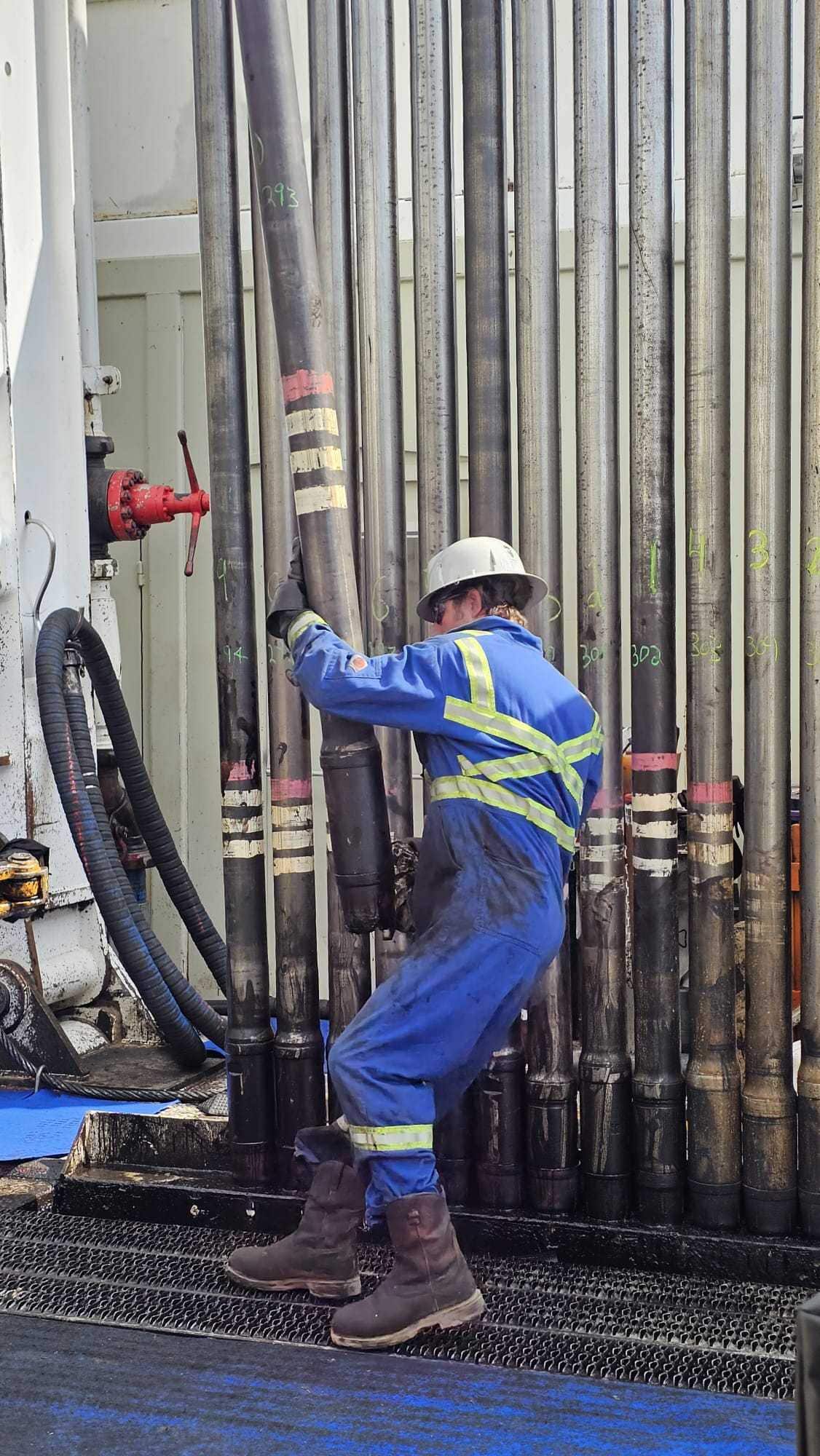 A man in blue safety gear wrangles an oil pipe in front of industrial equipment.