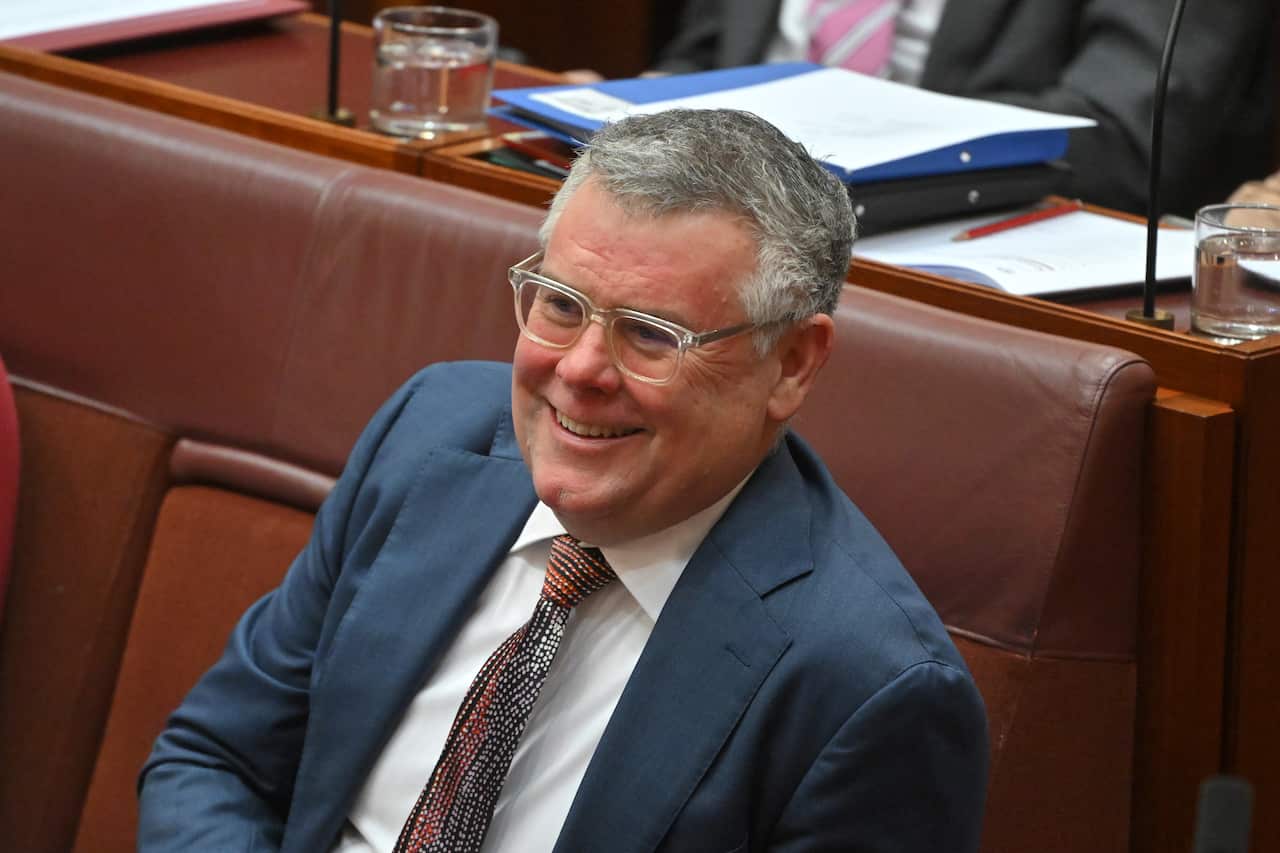 A man in a blue jacket, white shirt and dotted tie wearing glasses. He has short grey hair and is sitting on a red seat smiling.