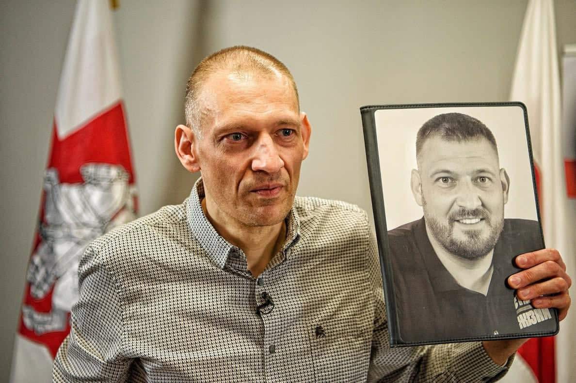 A thin man with a shaved head and a collared shirt holds up a framed portrait of a younger version of himself 