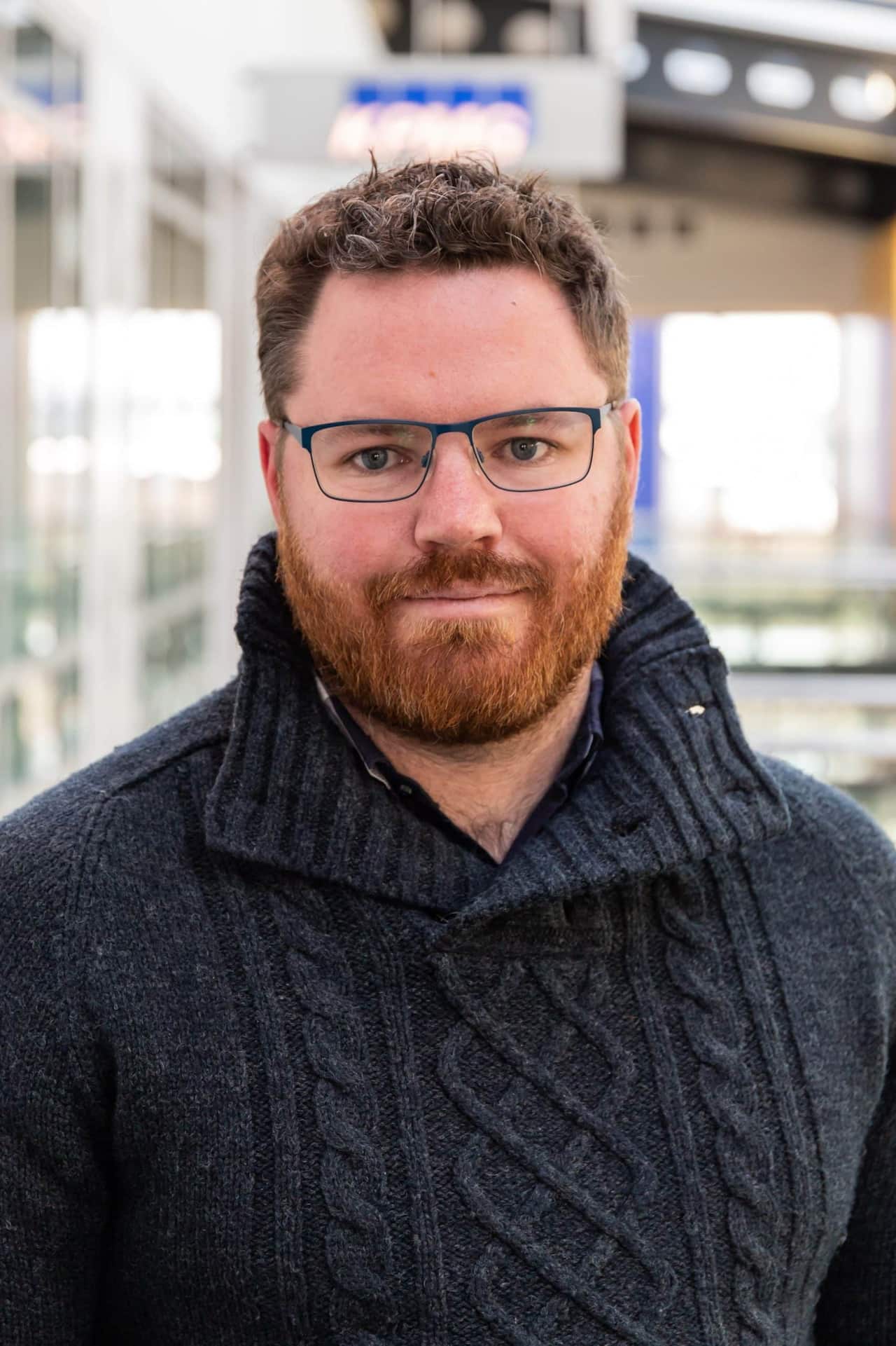 A man in glasses and a beard is wearing a woollen jumper as he poses.