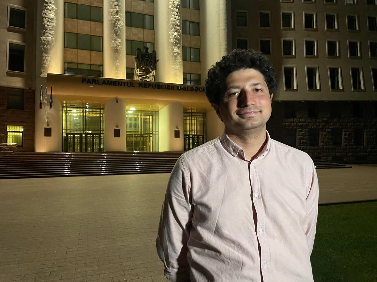 Man with curly dark hair standing in front of a building and steps. 