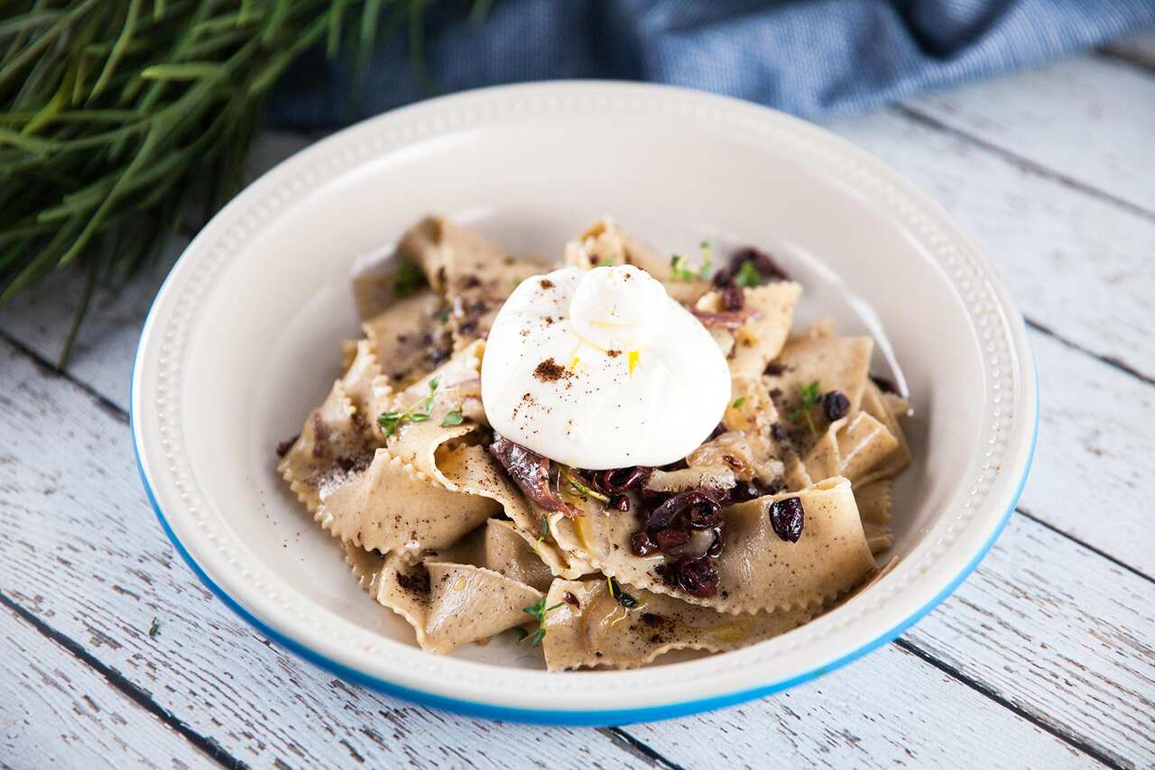 A white bowl with a blue rim sits on a white-washed wooden surface. The bowl holds ribbons of pasta, topped with a round of mozzarella. 