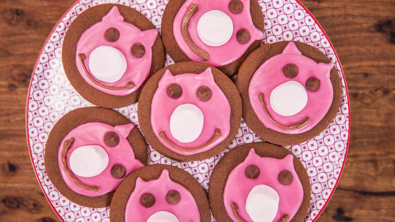 Seven round, flat chocolate biscuits, decorated with icing to form pink pig's faces, sit on a patterned plate.