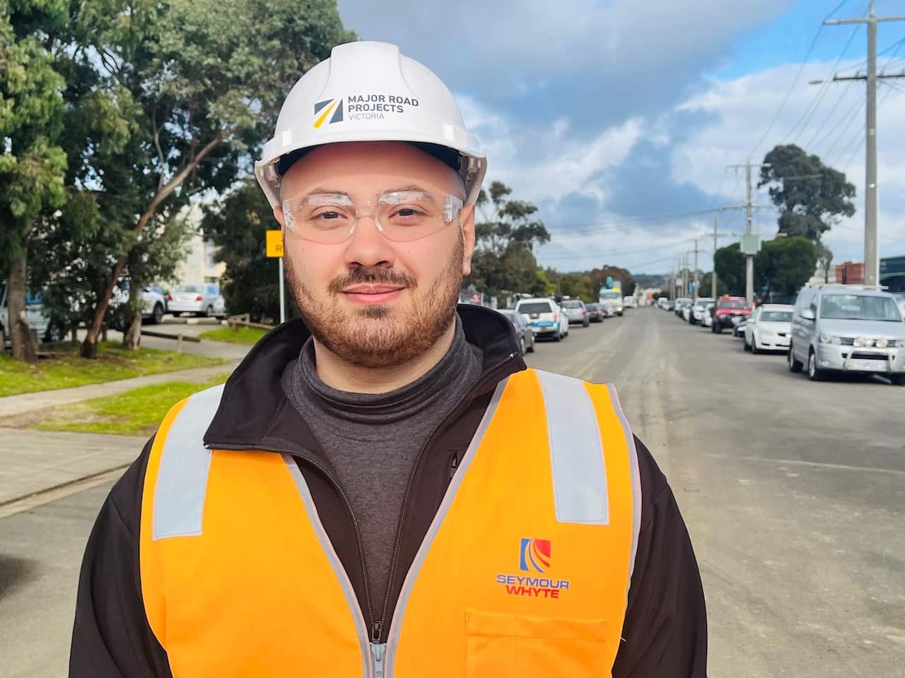 A man wearing an orange hi-vis vest, clear safety goggles and a white hard hat, standing outside.