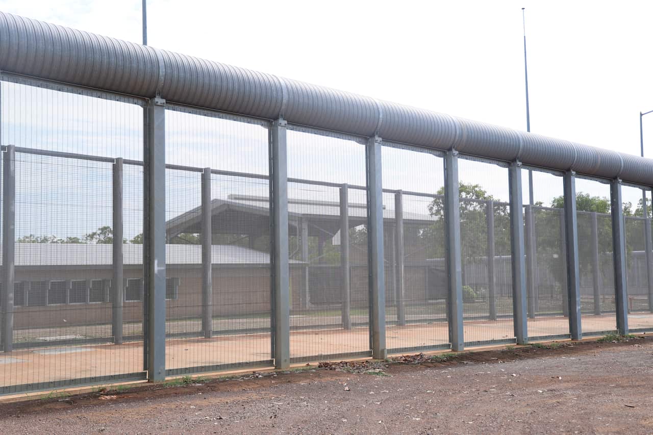 A screen fence with a building fringed by red dust and desert-region foliage