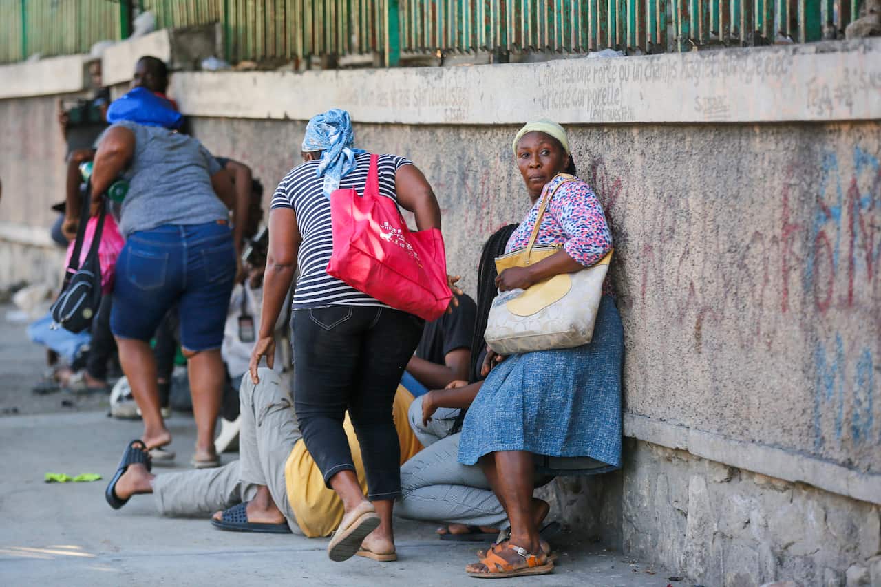 A group of civilians, mainly women, take shelter against a wall. A couple appear to be running for cover.