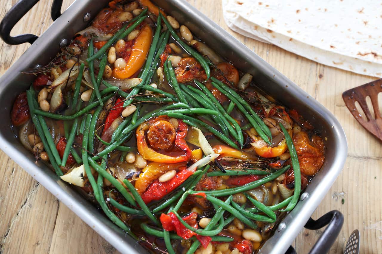A silver baking tray with black handles holds a bright vegetable stew, with long green beans and red capsicum pieces adding to the colour.  The edge of a stack of flatbreads can be seen alongside. 
