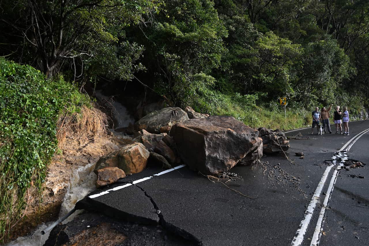 People standing next to a large rock that has fallen on a damaged road.