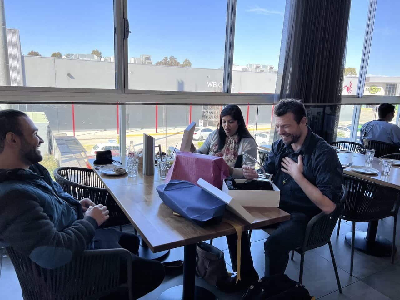 Two people open gifts on a restaurant table with surprised facial expressions. A man sits across from them, smiling. 