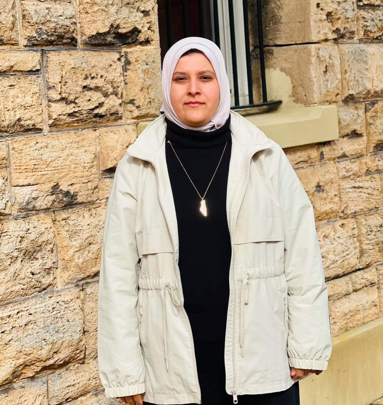 A medium shot of a woman wearing a light pink hijab, a black turtleneck, and an off-white jacket with a gold map necklace, standing outdoors in front of a textured sandstone wall.
