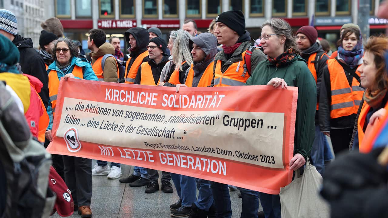 A group of men and women in winter clothing and orange safety vests are holding a banner in the German language