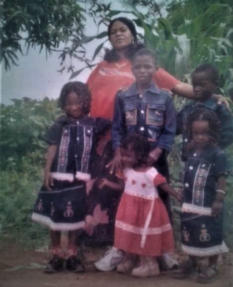 A group of children stand with a woman in a rural setting. 