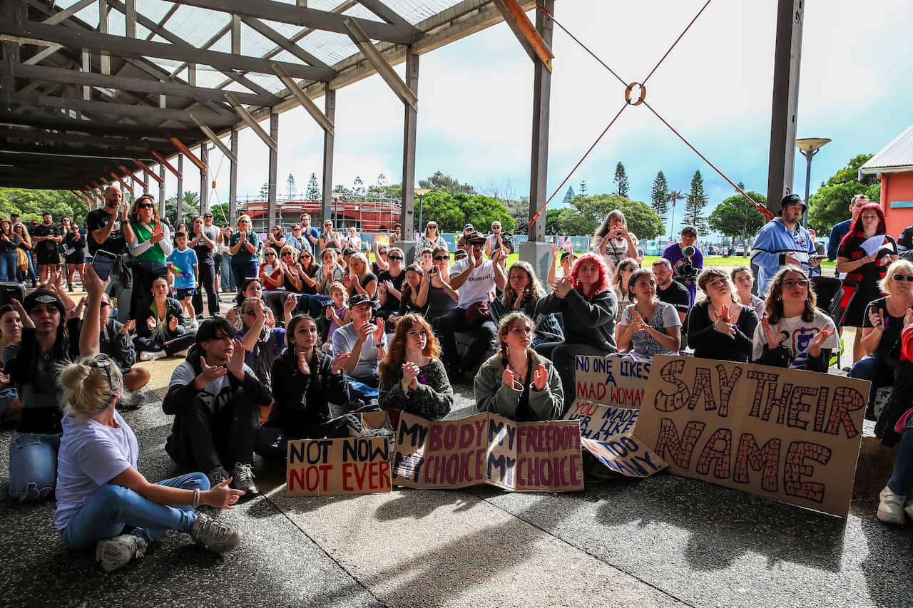 A crowd of people, both sitting and standing, hold signs during a protest against violence against women.