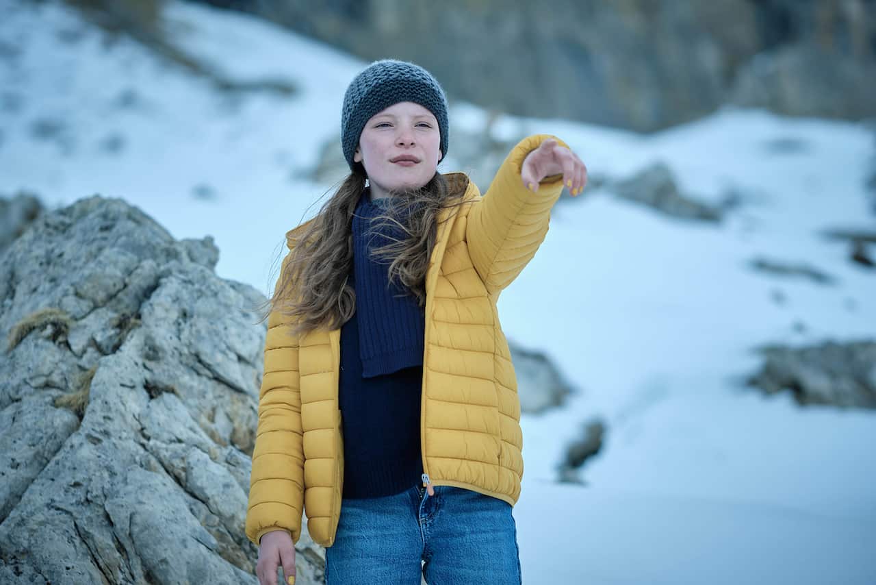 A girl in a beanie and bright yellow jacket stands in the snow, pointing.  
