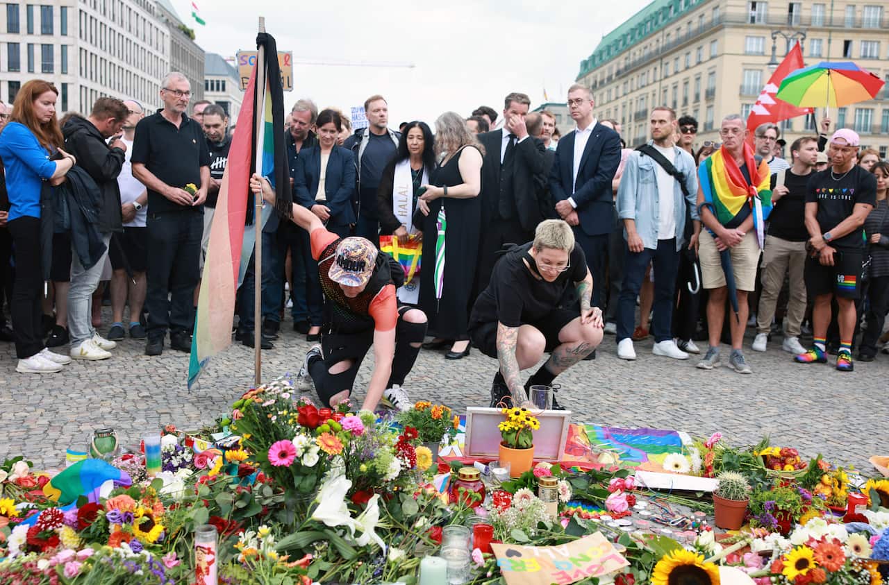 Two people lay flowers in a memorial with more people standing behind them, mourning.