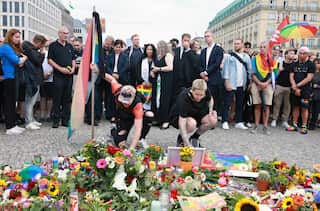 Two people lay flowers in a memorial with more people standing behind them, mourning.