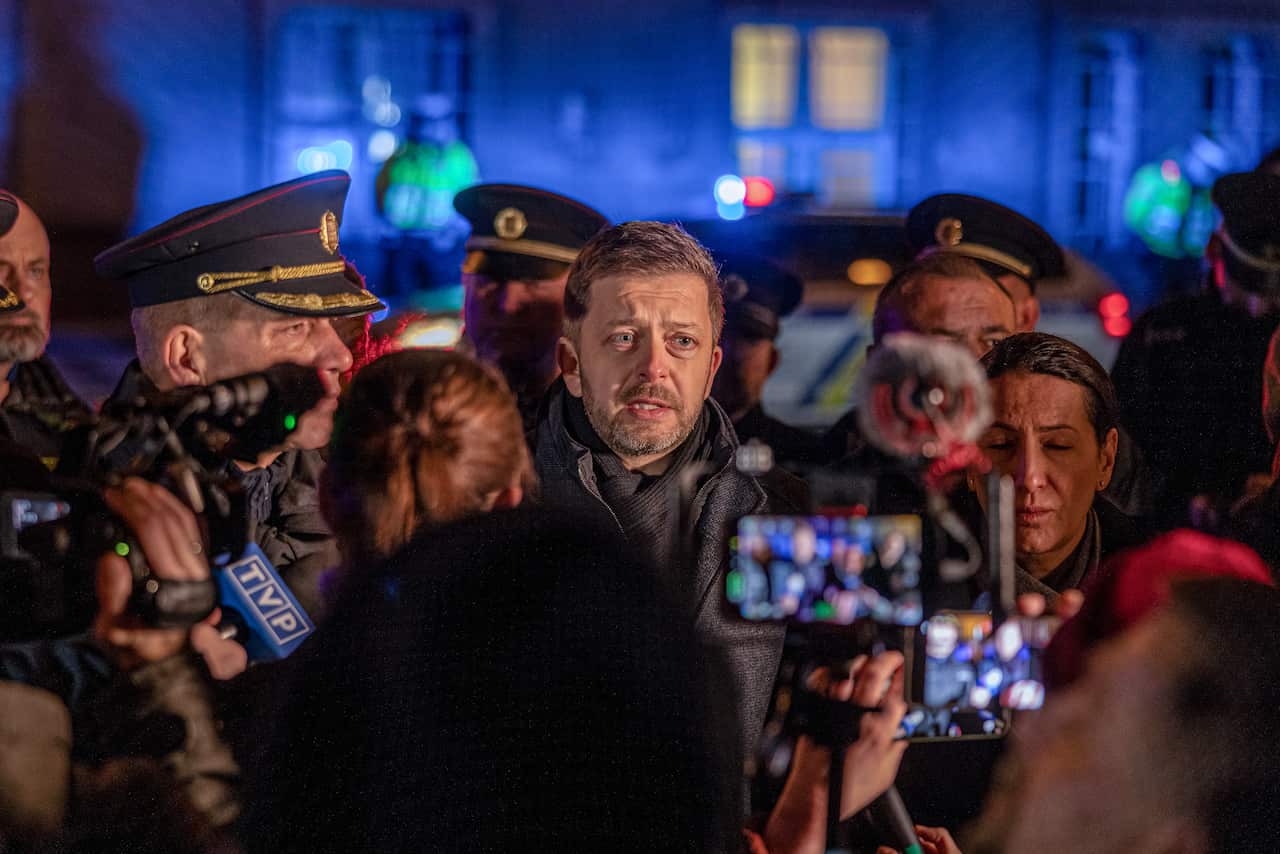 A middle-aged man surrounded by police officers addresses a press pack. 