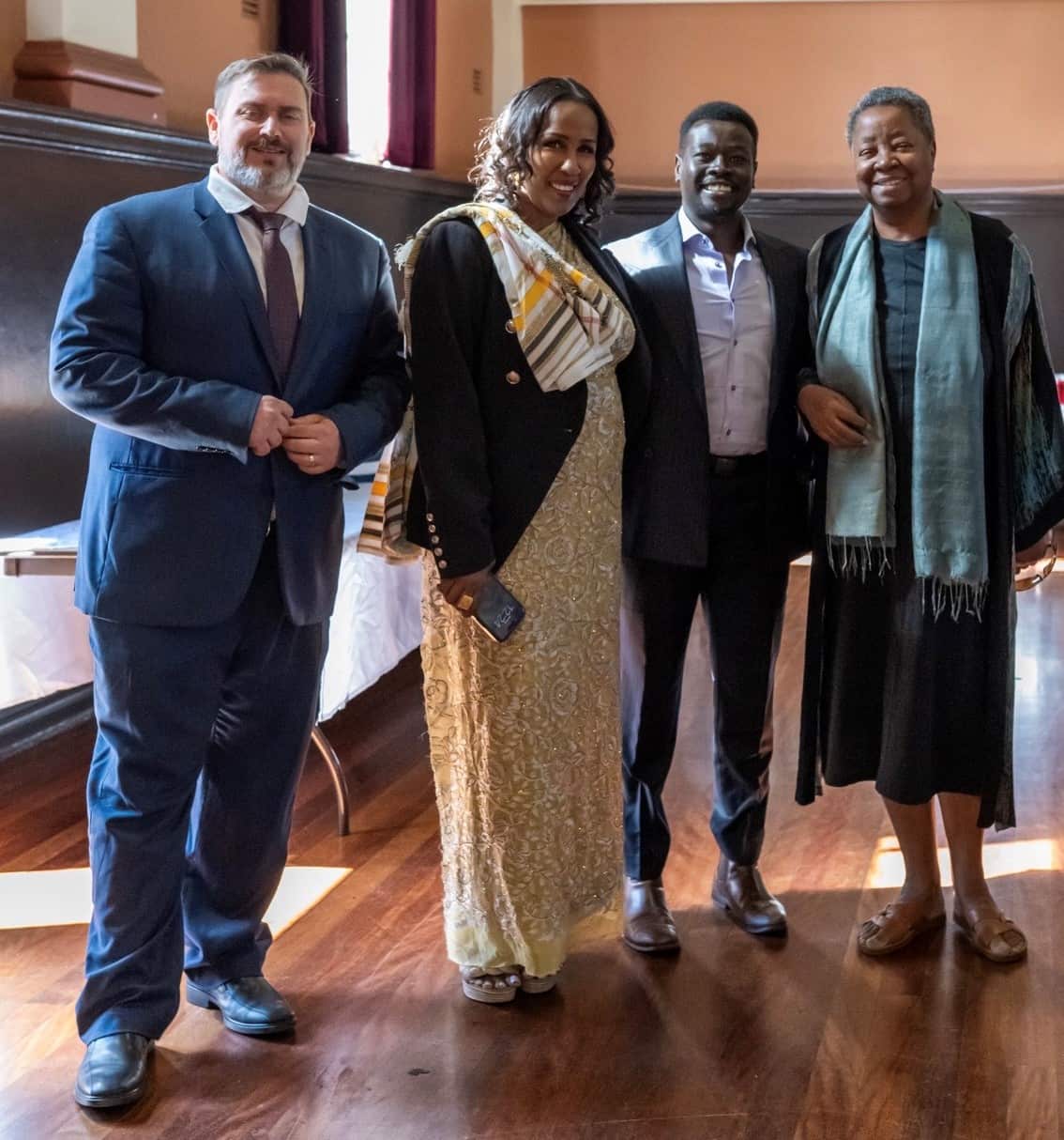 Four smiling individuals stand together indoors on a polished wooden floor, dressed in formal and traditional attire for an event.
