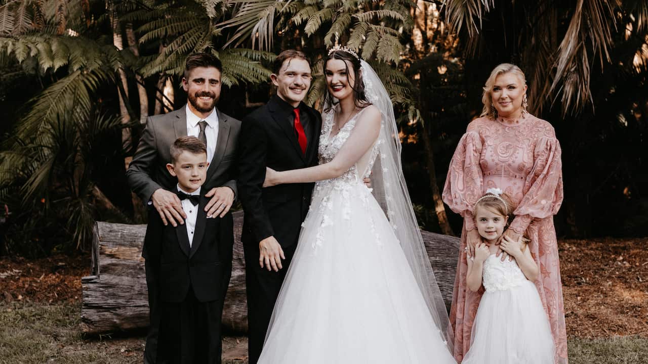 a portrait in a garden of a bride and a groom. A young man and young boy stand on the left of them and a young woman and young girl stand on the other side of the couple