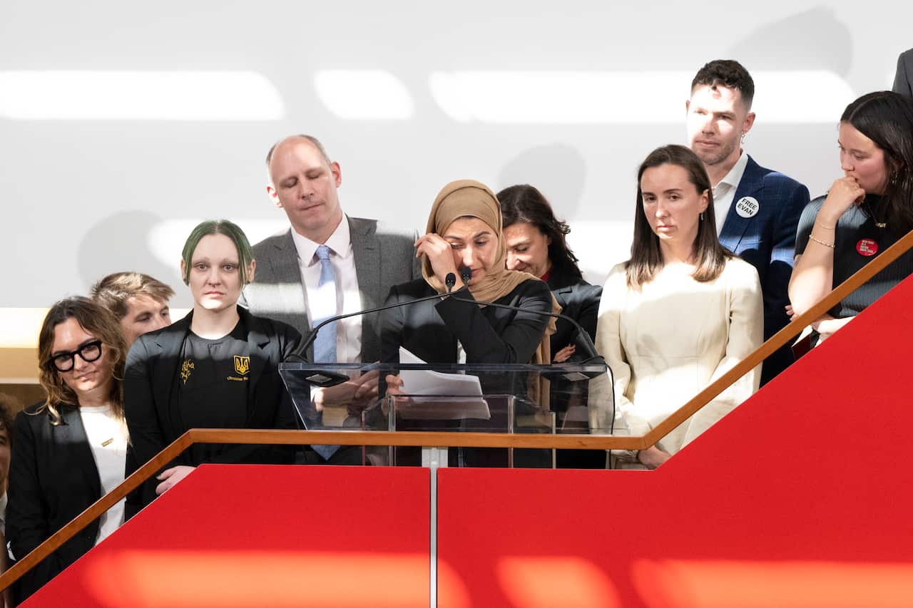 A woman in a black dress and beige hijab is brushing away a tear while standing at a lectern and holding notes in her right hand. She is surrounded by a group of men and women. 