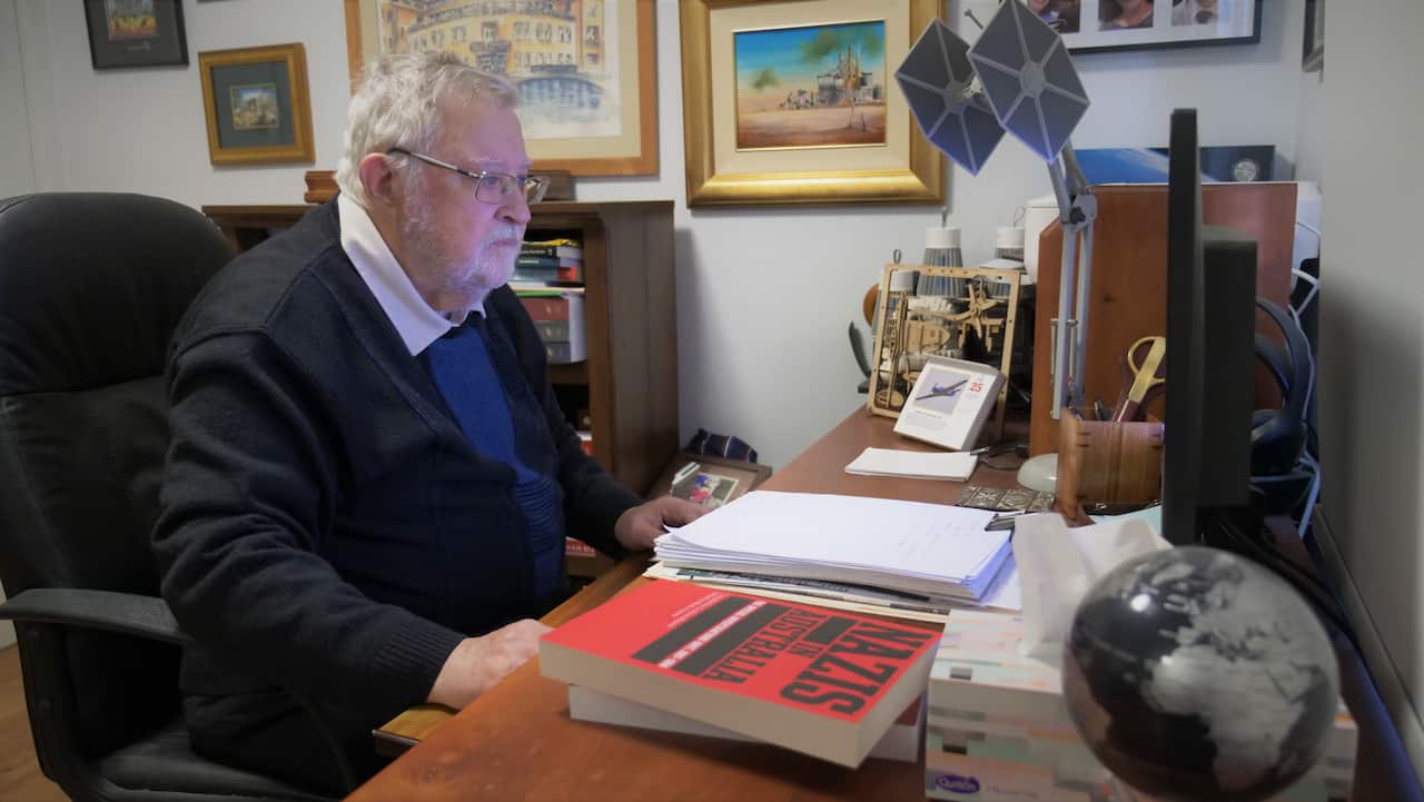 A middle-aged man sits at a desk, looking intently at a computer screen. On the desk beside him is a red book titled Nazis in Australia. Behind him, several artworks hang on the wall.