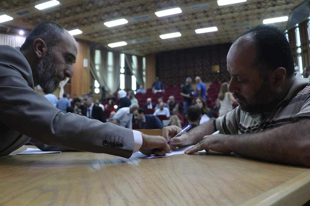 A man points to a paper another man is holding on a desk. There are people seated in the background