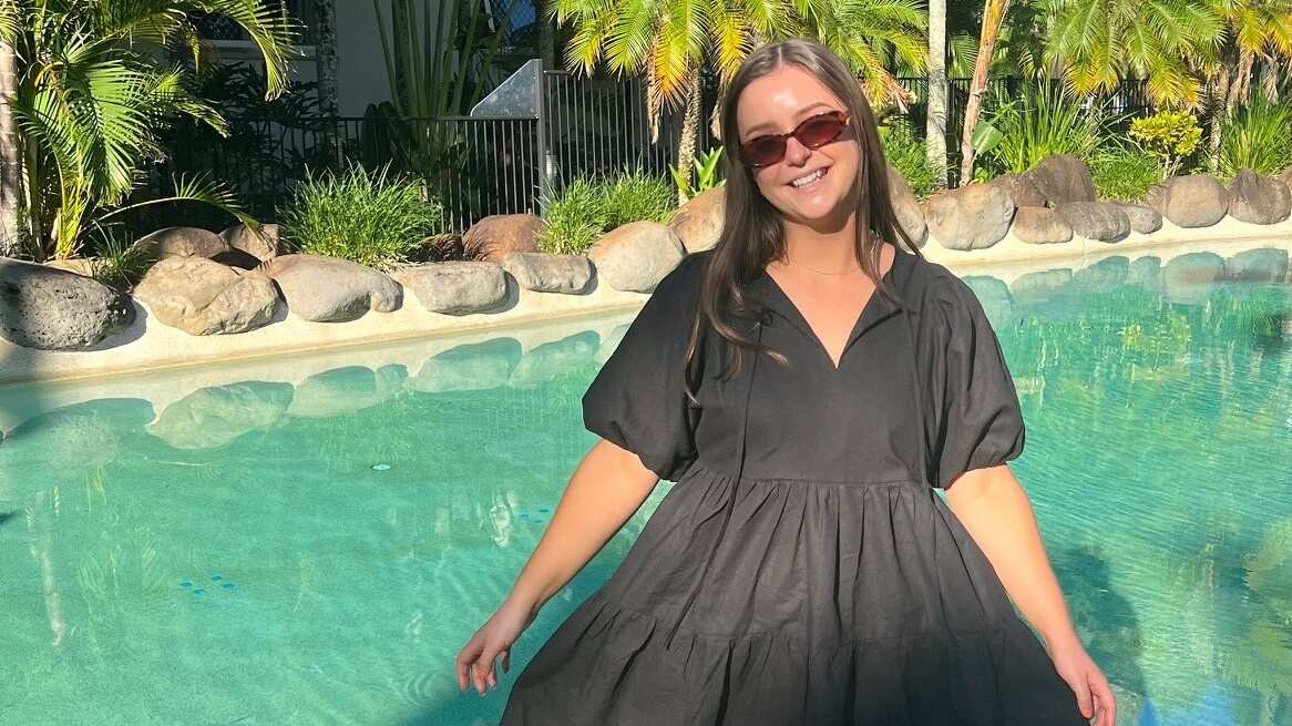 Young woman wearing a black dress and sunglasses standing outside in front of a pool and palm trees. 