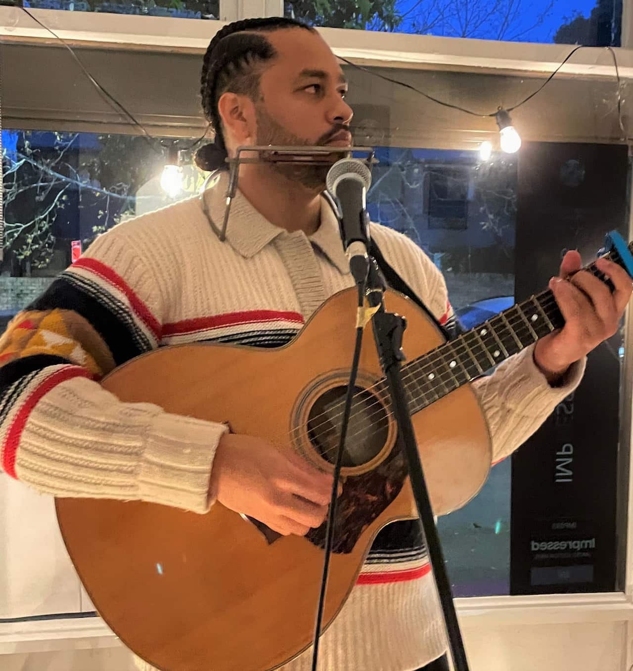 A man with braids and a white jumper holds a guitar standing at a microphone with a harmonica holder up at his mouth. 