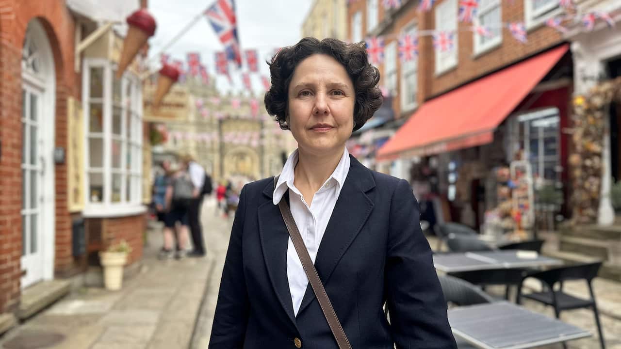 A middle-aged woman with short wavy hair, wearing a white shirt and black suit, poses for a photo on a street lined with buildings adorned with British flags