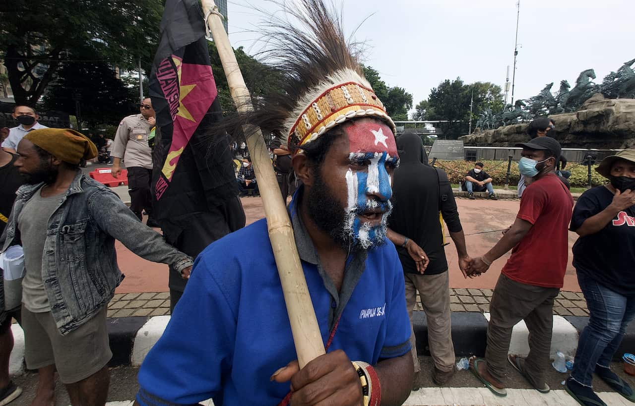 A man with the West Papuan flag painted on his face wears a blue shirt and traditional headdress while holding a sign.