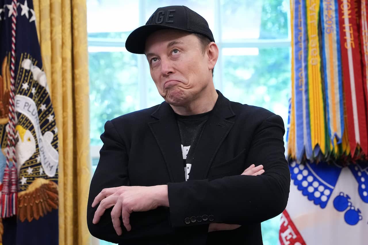 Elon Musk standing in the Oval Office. His arms are folded and he has a bemused expression.