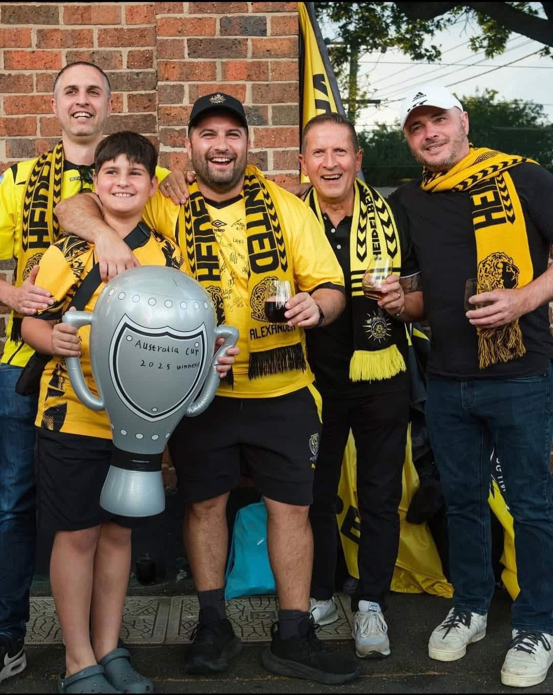 A group of men and a younger boy dressed in yellow scars and jerseys posing. The boy is holding a blow-up grey soccer trophy which says Australia Cup on it. 