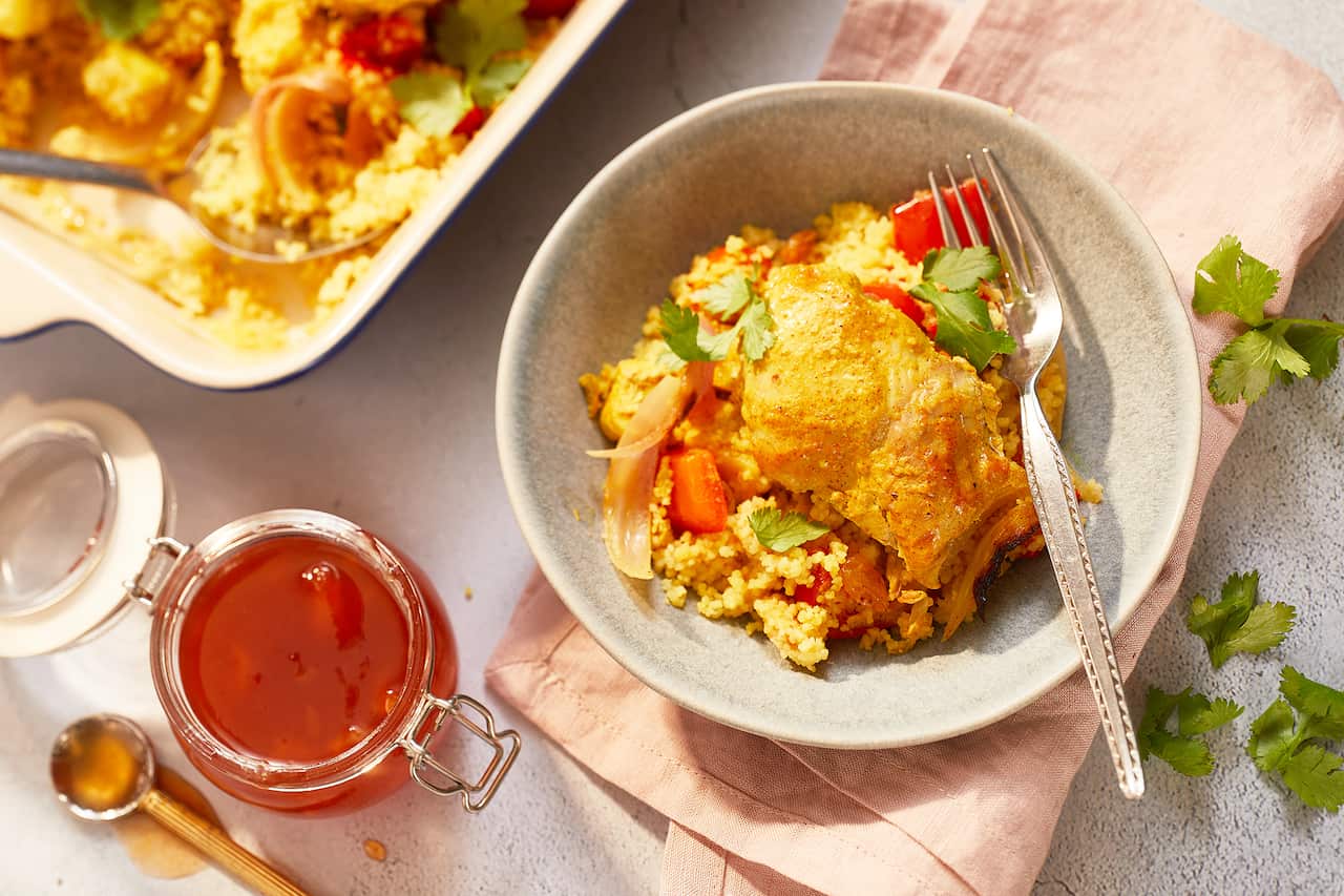 A speckled bowl, sitting on a pin napkin on a textured surface, holds a serve of a bright chicken and couscous dish. A 