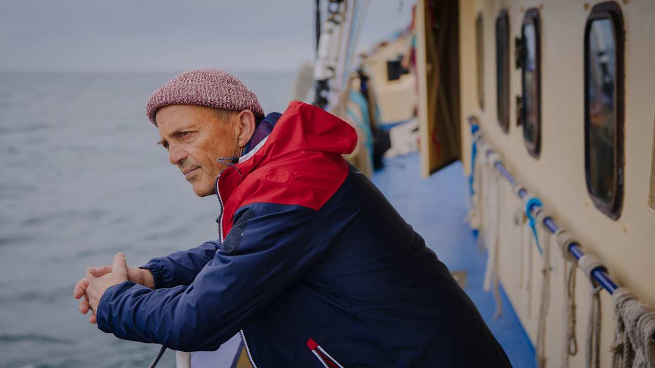 A man in a dark jacket leans on the railing of a boat at sea, looking pensively ahead. 