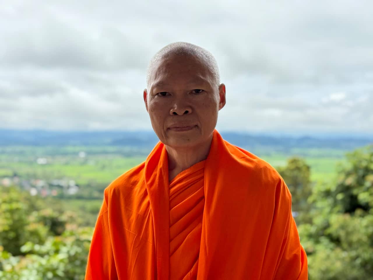 A monk in orange robes standing with a vista of wide, flat, green land behind him